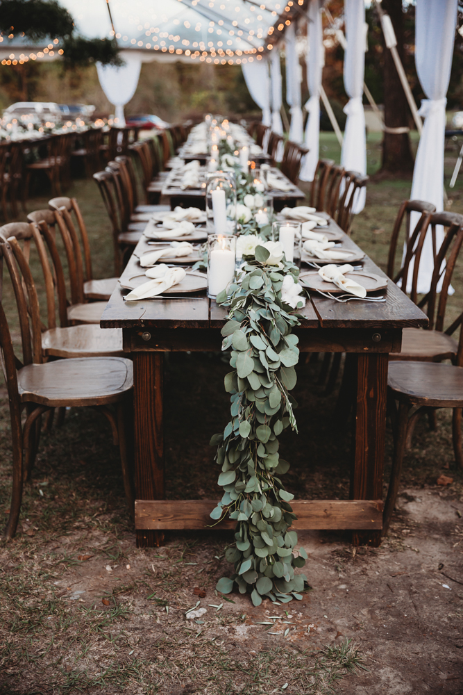 Long wooden reception table with cream place settings, eucalyptus garland, and glowing white candles under string lights