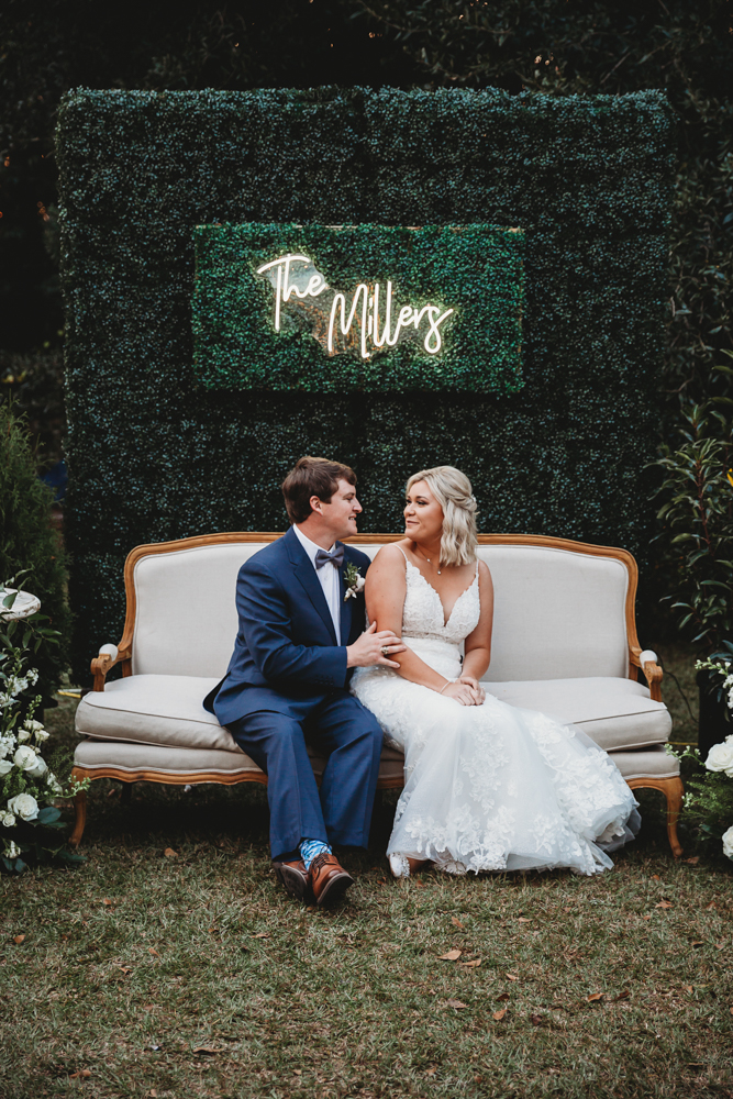 Bride and groom seated on cream couch in front of 'The Millers' neon boxwood wall during their reception at Wavering Place