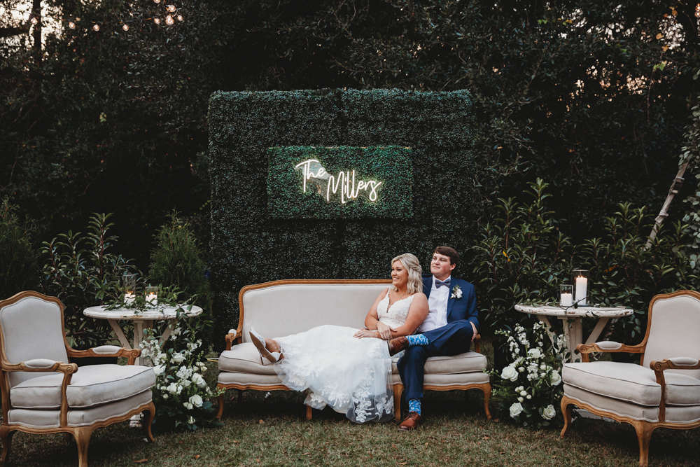 Bride and groom seated on cream couch in front of 'The Millers' neon boxwood wall during their reception at Wavering Place