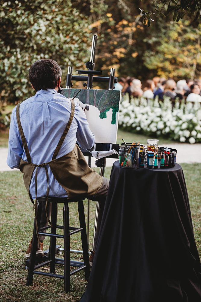 Live painter captures the wedding ceremony beneath mossy oaks with white florals and guests looking on at Wavering Place