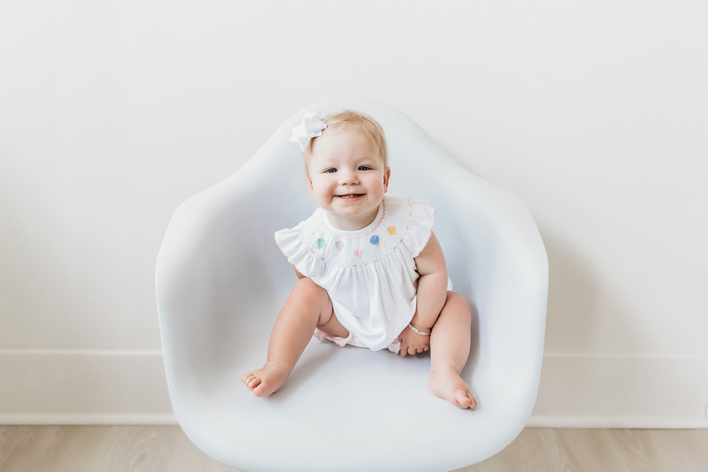 Hadley smiling in a white smocked romper with balloon embroidery and white bow, seated in a modern white chair at Reflection Images by Tracy Rowell in Florence SC