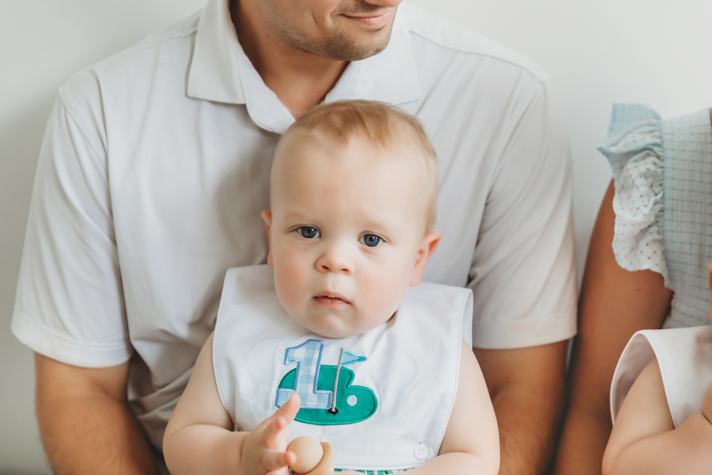 Close-up of one twin sitting on dad's lap wearing a white 1b bib at Reflection Images by Tracy Rowell in Florence SC