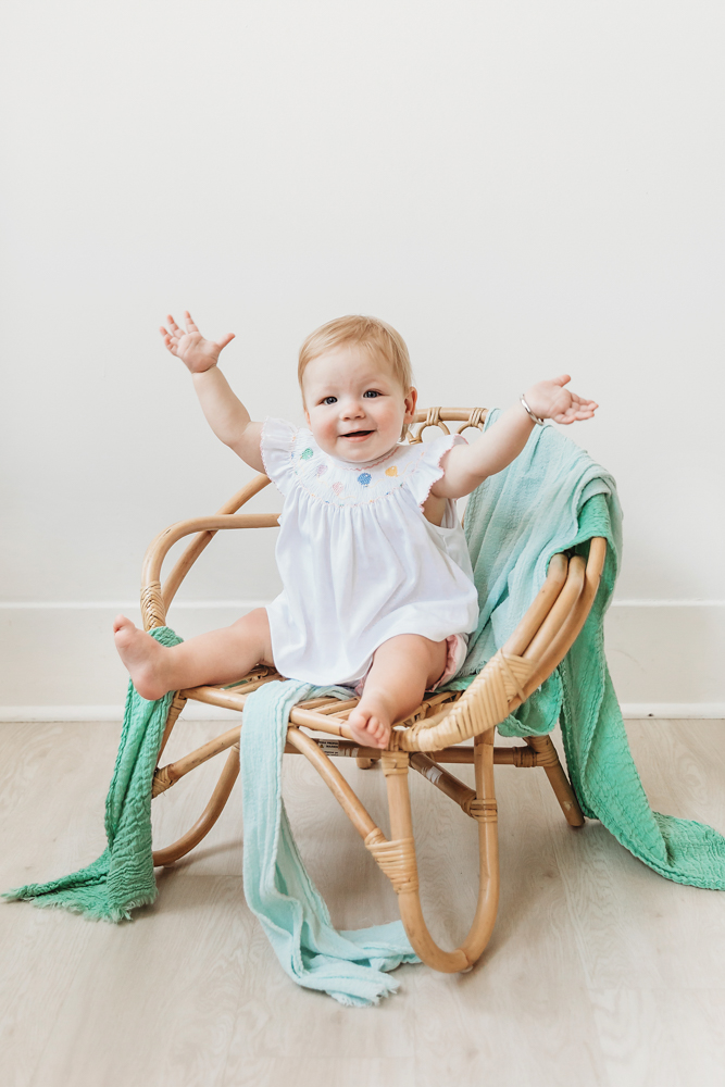 Hadley in a wicker chair with aqua blankets, wearing white romper, arms raised and smiling big at the studio