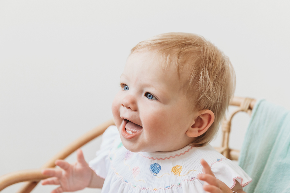Hadley in a wicker chair with aqua blankets, wearing white romper, arms raised and smiling big at the studio