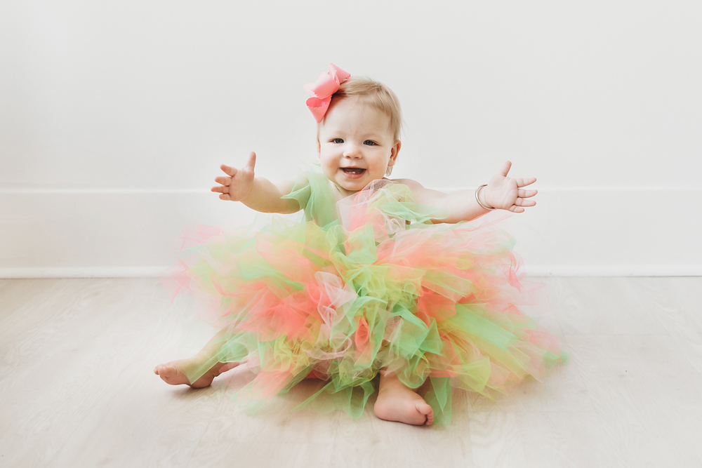 Hadley reaching out in a rainbow tutu dress with a big smile, seated against a white wall in the studio