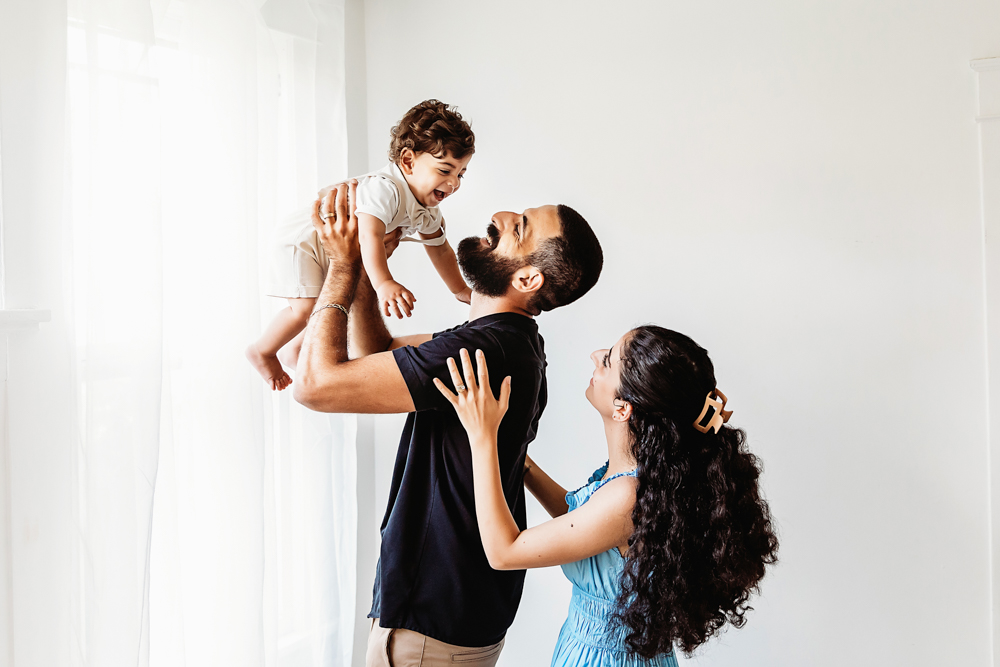 Mom watches as dad lifts smiling Lucas near sheer window curtains in a bright studio at Reflection Images by Tracy Rowell in Florence SC