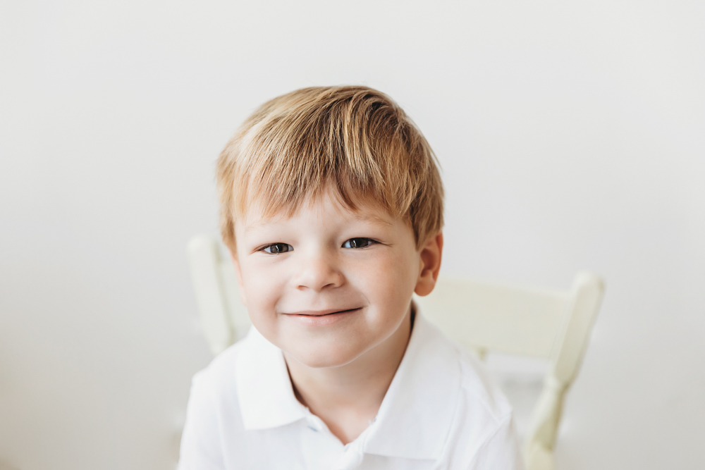Portrait of Hadley's big brother in a white polo shirt, smiling with bright window light at Reflection Images