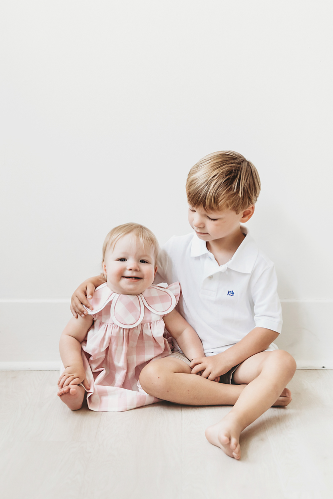 Hadley in a pink plaid dress sitting beside her big brother, arm around her, on a light wood floor at Reflection Images by Tracy Rowell in Florence SC