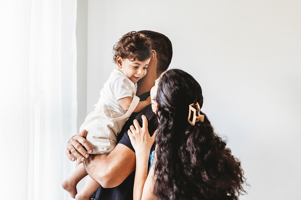 Lucas smiles over dads shoulder as mom reaches toward him in bright window light at Reflection Images by Tracy Rowell in Florence SC