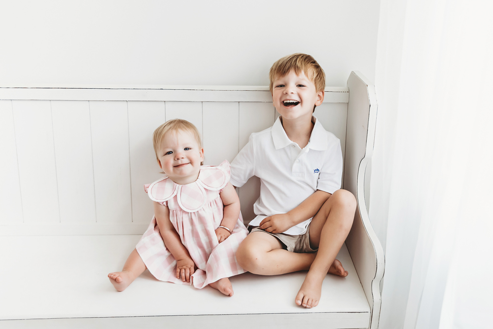 Hadley and her brother laughing together on a white bench with soft window light at the studio