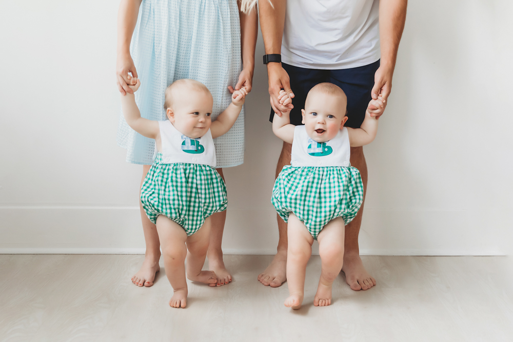 Twins standing while holding hands with parents, showing off 1b bibs and gingham outfits at Reflection Images by Tracy Rowell in Florence SC