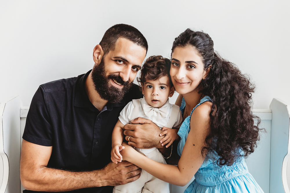 Lucas sits centered between mom and dad on a white bench during a soft natural light family portrait at Reflection Images by Tracy Rowell in Florence SC