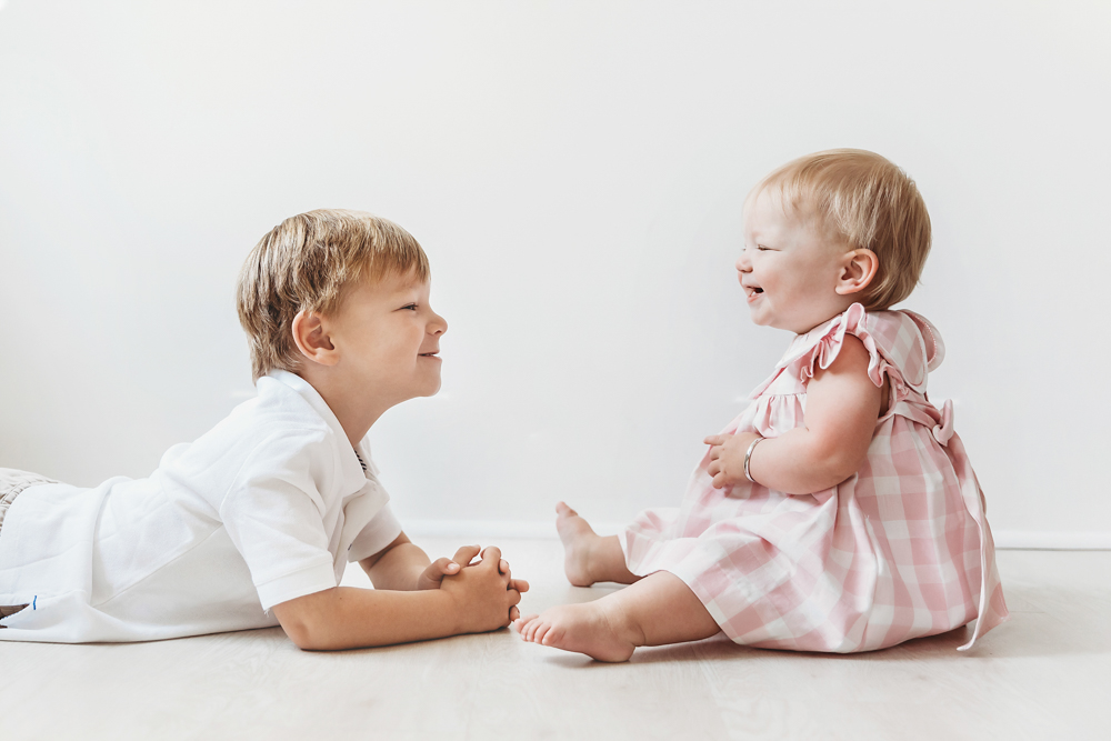 Hadley and her brother giggling on the floor, nose-to-nose with smiles, white wall background