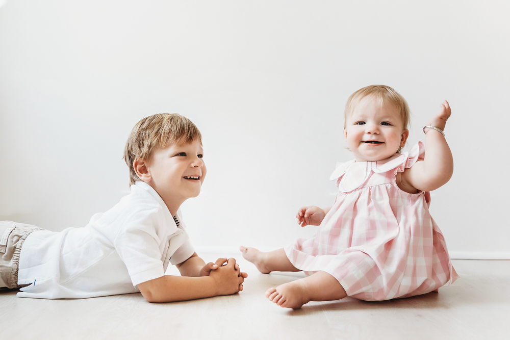 Hadley waving beside her smiling brother, both sitting on the studio floor with clean white backdrop
