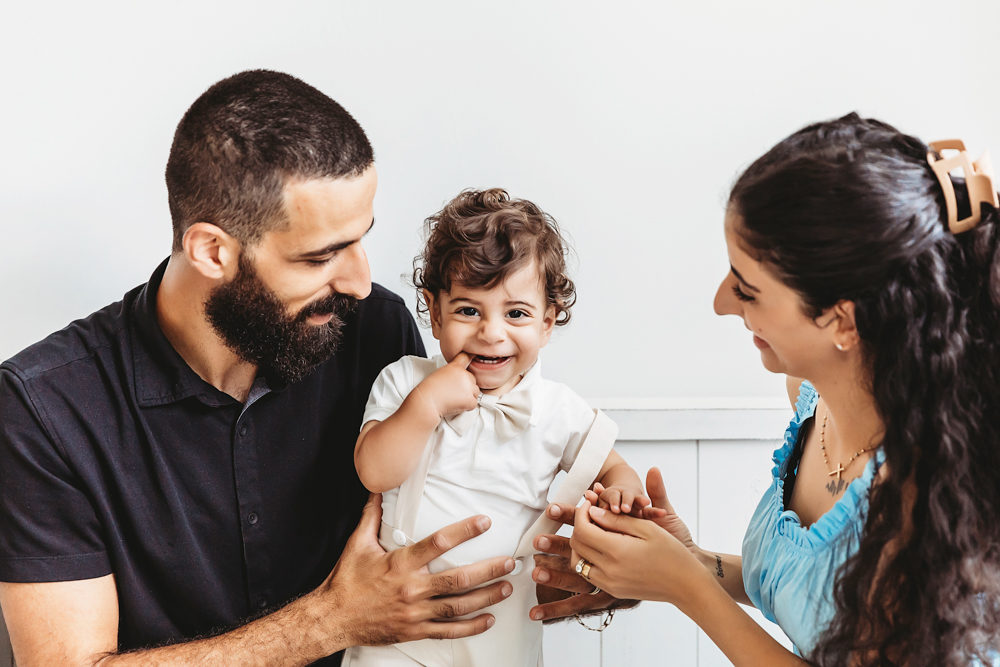 Lucas smiles brightly while seated between his parents on a white bench in airy studio light at Reflection Images by Tracy Rowell in Florence SC