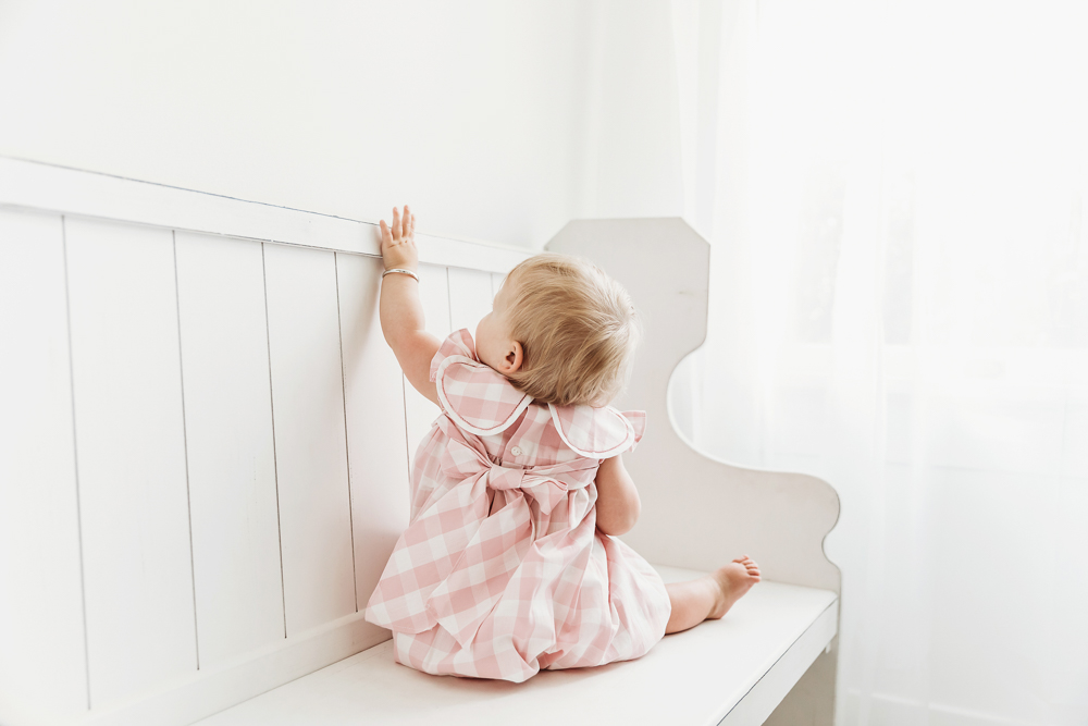Back view of Hadley reaching toward the top of a white bench, showing dress bow and soft window light