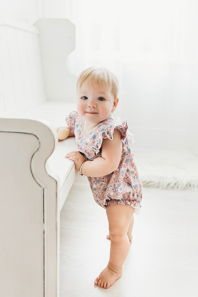 Hadley standing beside a white studio bench in a floral romper, holding a macaron and gazing sweetly