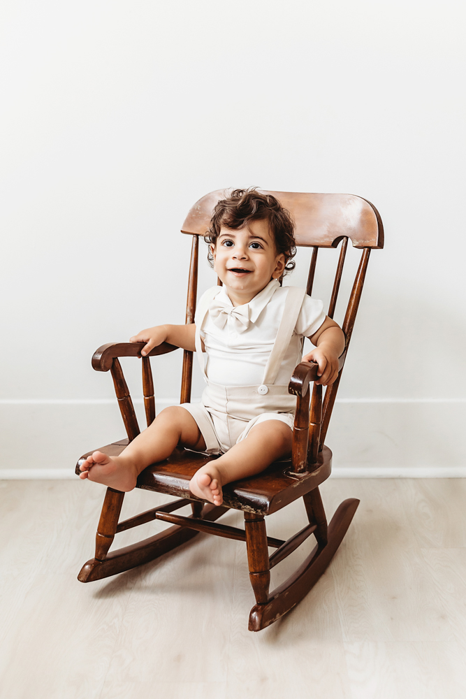 Lucas sitting in a vintage wooden rocking chair wearing a cream outfit in soft studio light at Reflection Images by Tracy Rowell in Florence SC