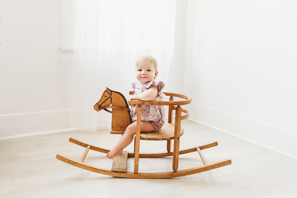 Hadley smiling while seated on a vintage wooden rocking horse with soft white curtains behind her