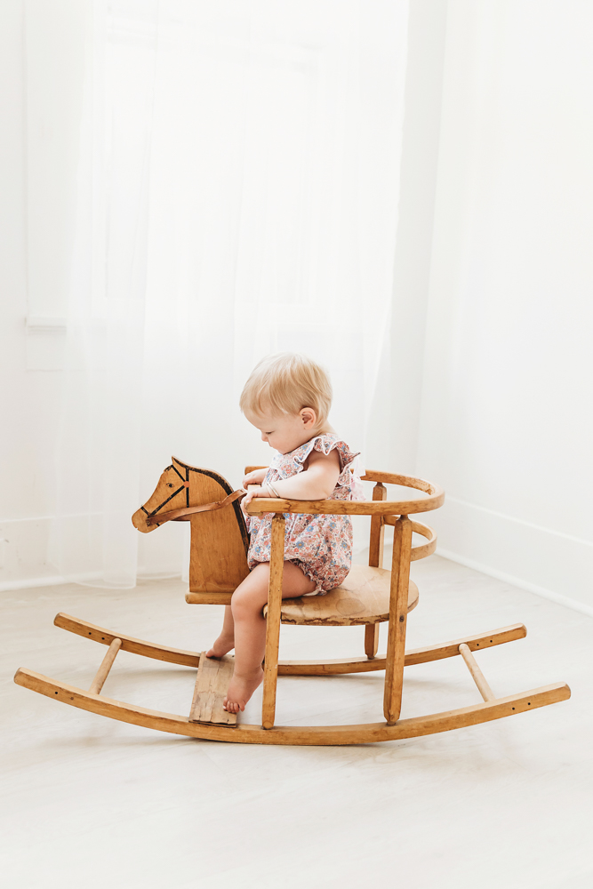 Hadley looking down while riding a wooden rocking horse, floral romper and window light in the studio
