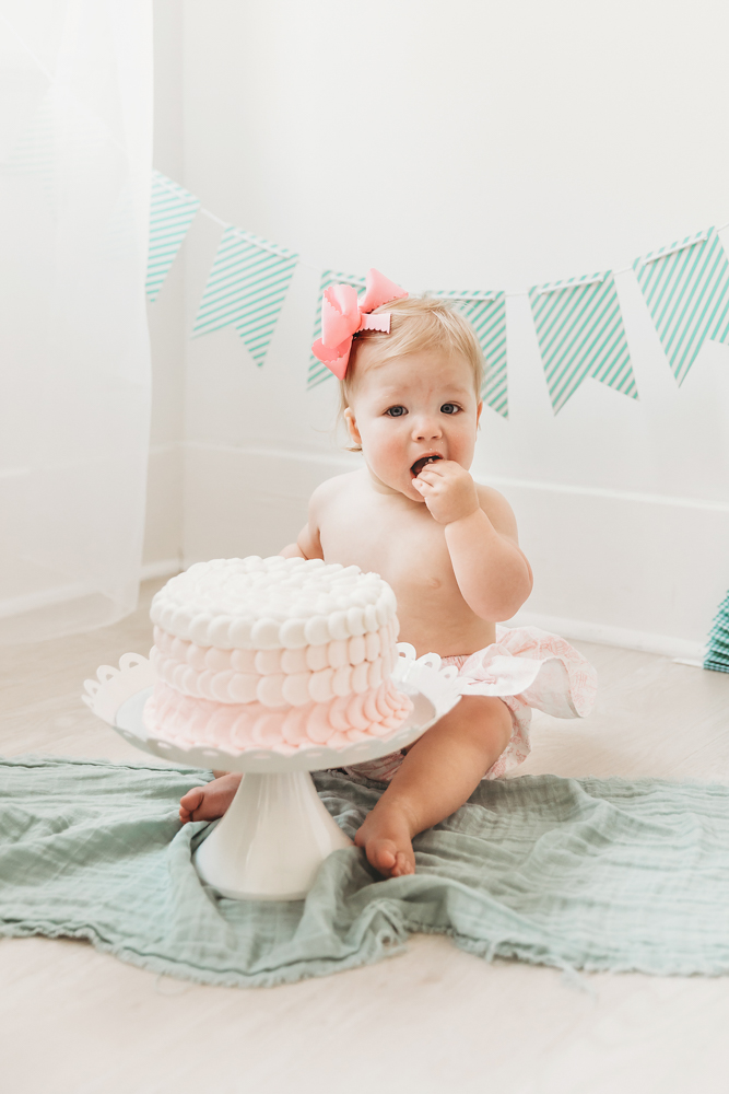 Hadley beginning her cake smash, wearing pink bloomers and bow, in front of green-striped bunting
