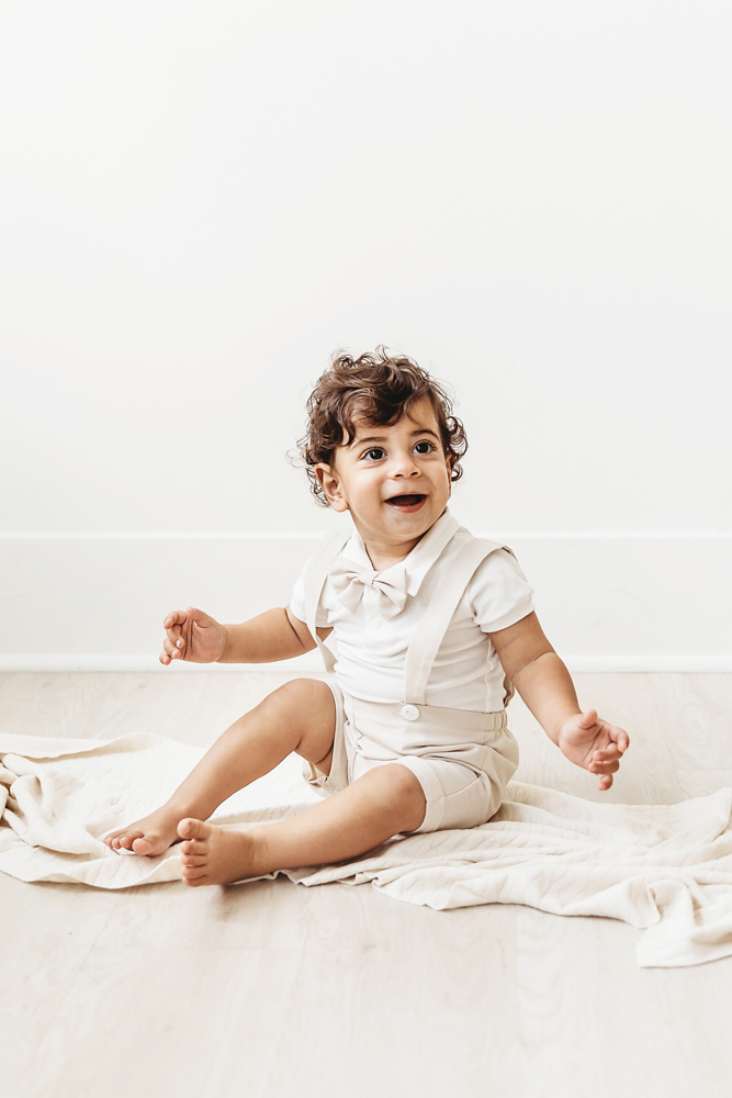Lucas sitting on a cream blanket wearing a bow tie romper and smiling on the studio floor at Reflection Images by Tracy Rowell in Florence SC