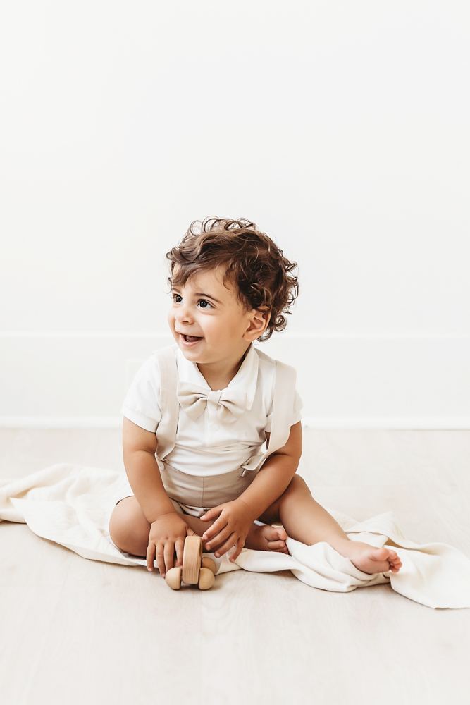 Lucas holds a wooden toy car while sitting on a cream blanket and smiling in soft bright studio light at Reflection Images by Tracy Rowell in Florence SC