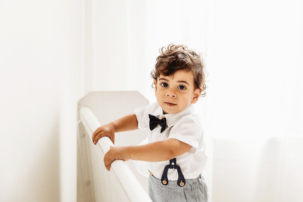 Lucas stands holding a crib rail wearing a white shirt and dark bow tie in bright window light at Reflection Images by Tracy Rowell in Florence SC