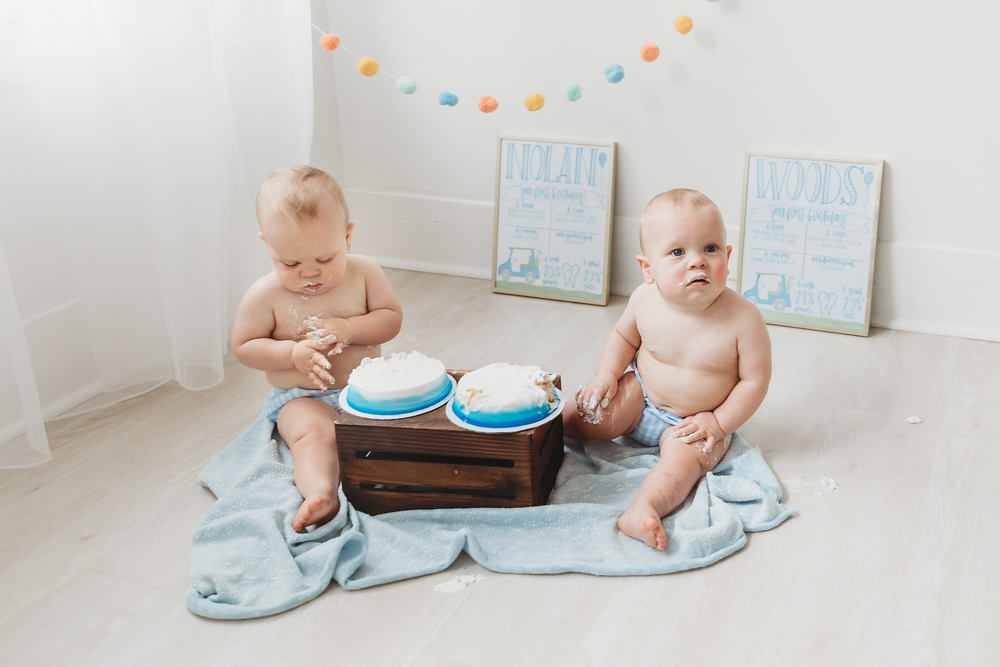 One twin eats cake while the other looks up, surrounded by cake and milestone signs during a cake smash at Reflection Images by Tracy Rowell in Florence SC