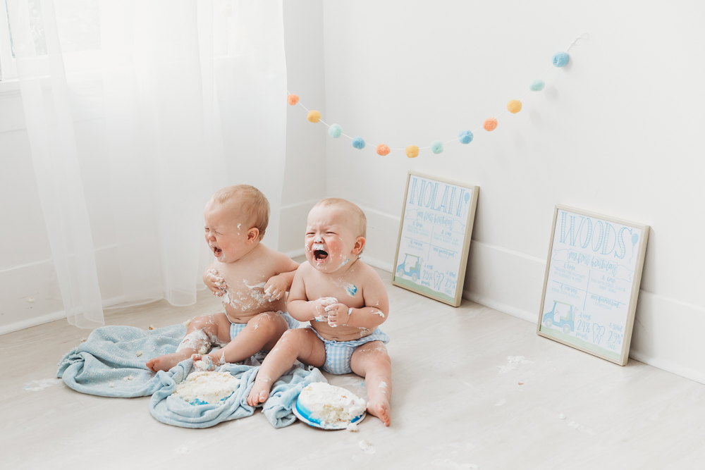 Both twins crying mid-cake smash, covered in frosting and surrounded by cake crumbs at Reflection Images by Tracy Rowell in Florence SC