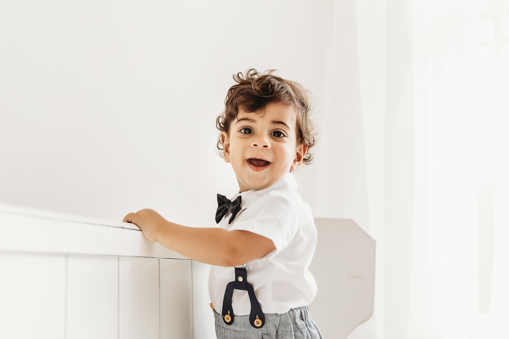 Lucas smiles while standing at a white crib rail wearing suspenders and bow tie in airy window light at Reflection Images by Tracy Rowell in Florence SC