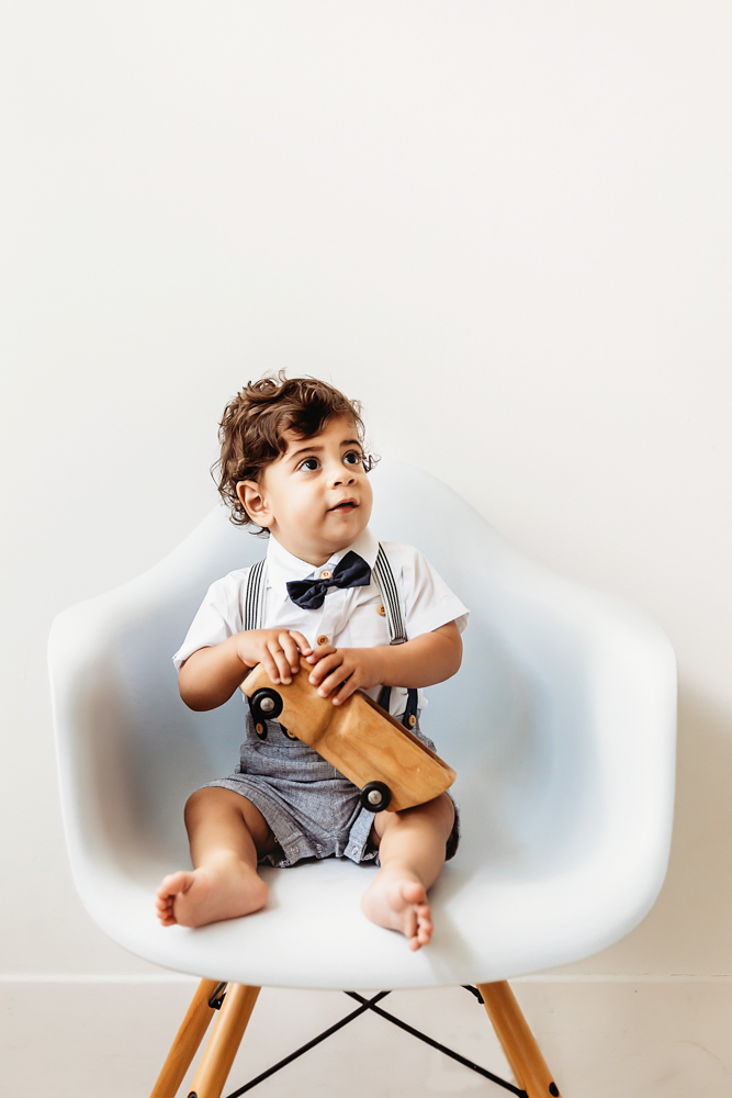 Lucas sits in a modern white chair holding a wooden toy truck while looking upward in soft natural light at Reflection Images by Tracy Rowell in Florence SC