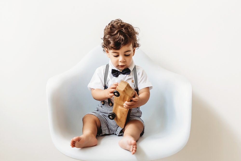 Lucas sits barefoot in a white chair closely examining a wooden truck toy against a clean white backdrop at Reflection Images by Tracy Rowell in Florence SC