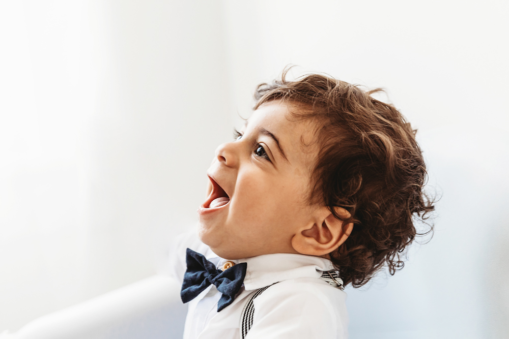 Closeup of Lucas laughing with mouth open wearing a navy bow tie beside soft window curtains at Reflection Images by Tracy Rowell in Florence SC