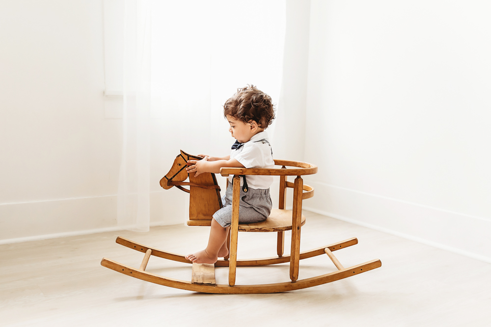 Lucas sits sideways on a vintage wooden rocking horse holding the handles in soft white studio light at Reflection Images by Tracy Rowell in Florence SC