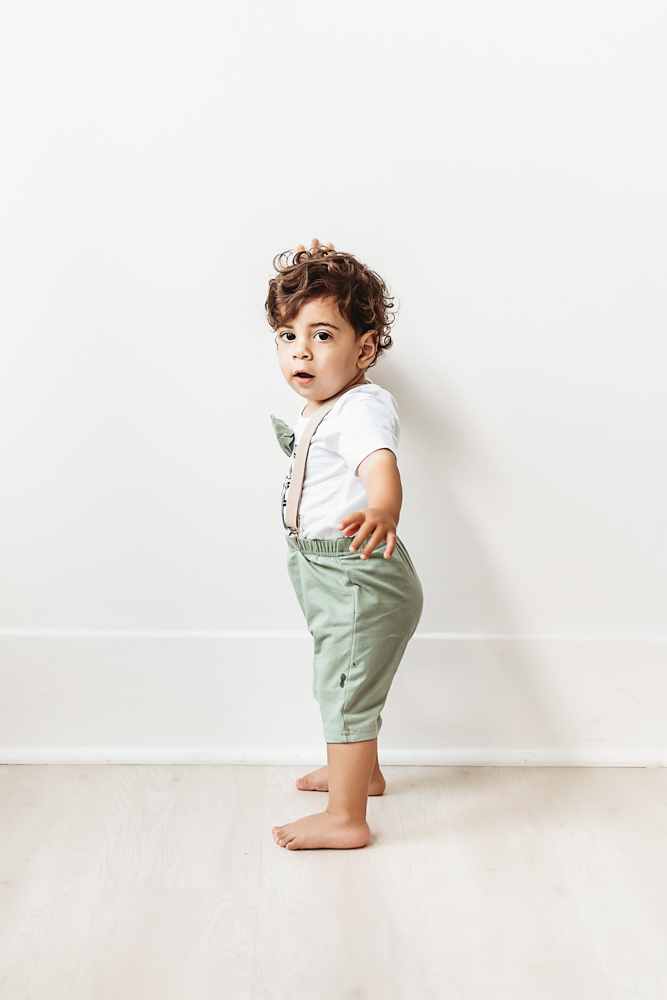Lucas stands against a clean white wall wearing green pants with suspenders and looking over his shoulder at Reflection Images by Tracy Rowell in Florence SC