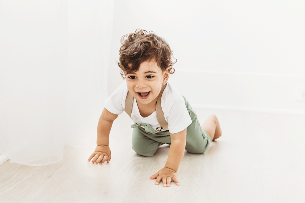 Lucas crawls across the bright studio floor laughing in a green outfit near soft white curtains at Reflection Images by Tracy Rowell in Florence SC