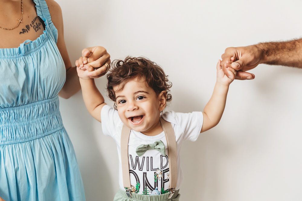 Lucas holds both parents hands while standing happily in his Wild One shirt against a clean white wall at Reflection Images by Tracy Rowell in Florence SC
