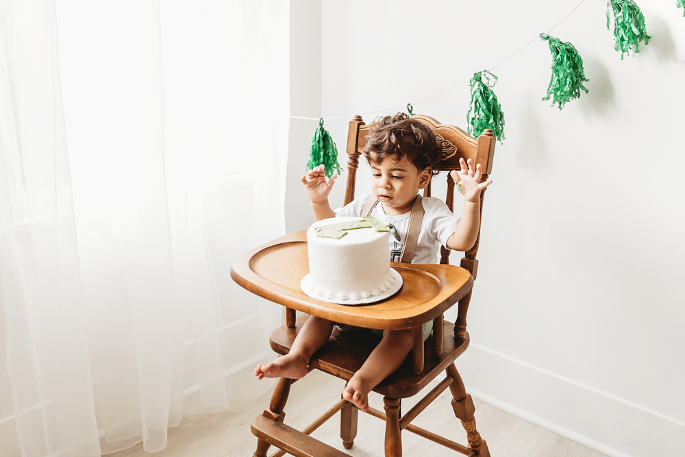 Lucas sits in a wooden highchair looking curiously at his birthday cake with green tassel banner behind him at Reflection Images by Tracy Rowell in Florence SC
