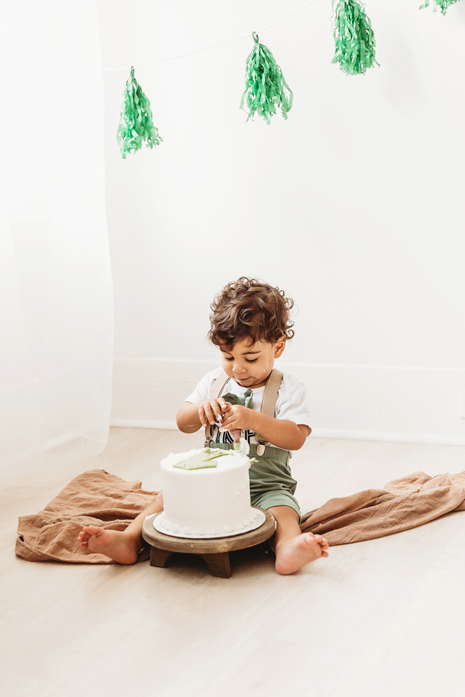 Lucas sits on the studio floor touching his birthday cake for the first time beneath hanging green tassels at Reflection Images by Tracy Rowell in Florence SC