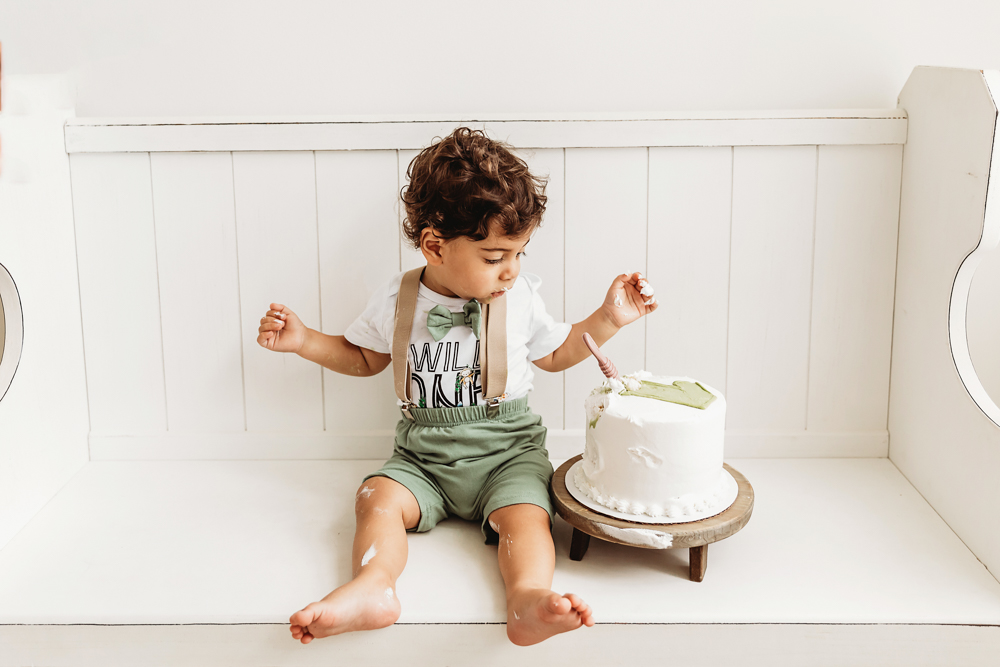 Lucas sits on a white bench with icing on his legs while examining his birthday cake in bright studio light at Reflection Images by Tracy Rowell in Florence SC