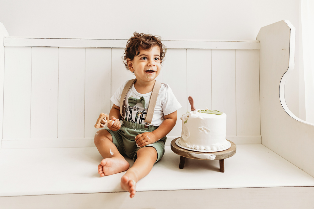 Lucas smiles with messy frosting covered hands while seated beside his cake on a white bench at Reflection Images by Tracy Rowell in Florence SC