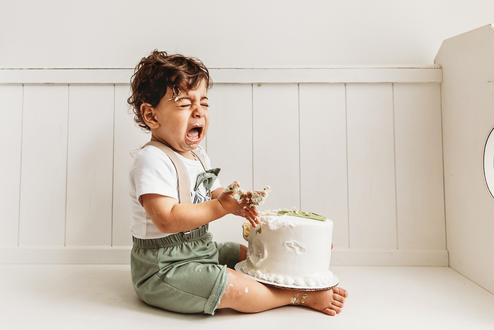 Lucas cries with frosting in his hands during a big cake smash reaction while seated on a white bench at Reflection Images by Tracy Rowell in Florence SC