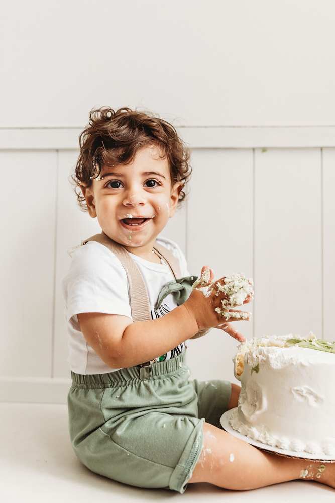 Lucas smiles with frosting on his cheeks while holding a handful of cake beside the smash cake at Reflection Images by Tracy Rowell in Florence SC