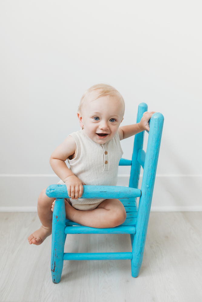 Smiling baby in cream knit romper sitting on turquoise wooden chair against white backdrop with soft natural lighting