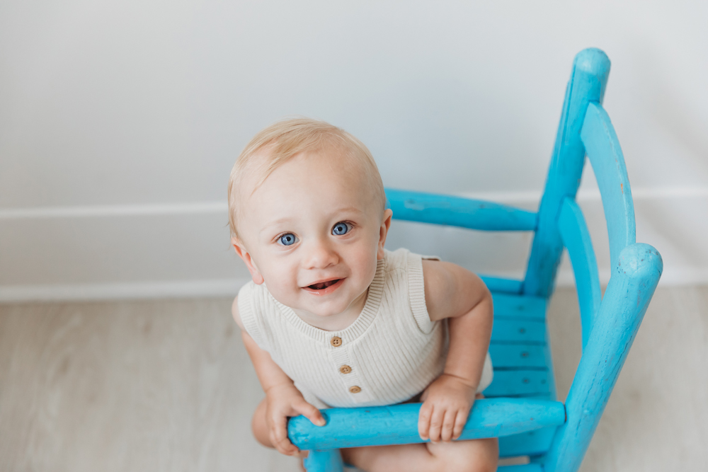 Smiling baby in cream knit vest sitting on bright blue wooden chair in professional photography studio with soft lighting