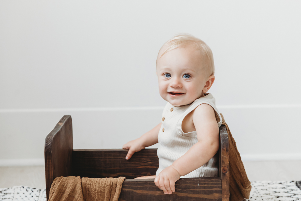 Smiling baby in cream knit romper sitting in rustic wooden crate against white wall backdrop with soft natural lighting