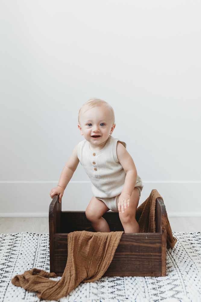 Smiling baby in cream ribbed romper sitting in wooden crate with brown blanket, soft lighting - Reflection Images by Tracy Rowell