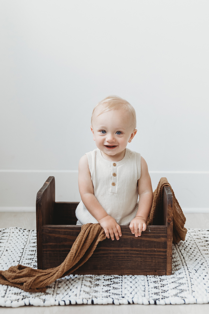 Smiling baby in cream ribbed romper sitting in wooden crate with brown blanket on white textured rug, soft studio lighting
