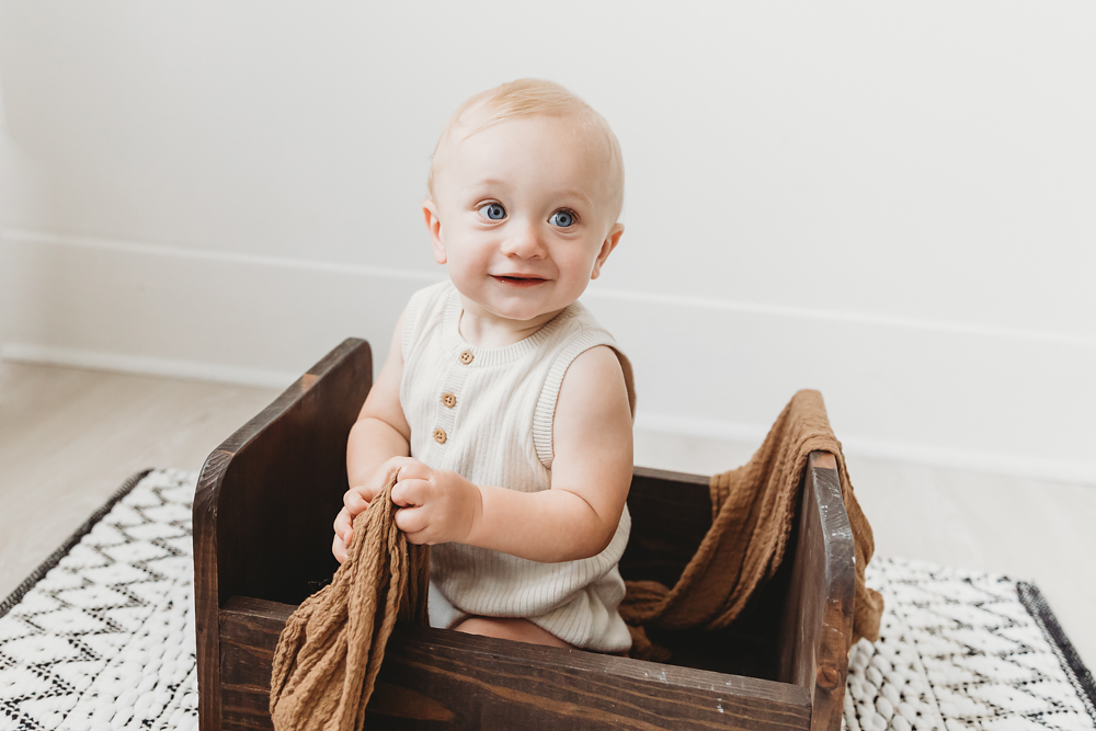 Smiling baby in cream knit romper sitting in wooden crate with brown blanket, studio portrait by Tracy Rowell